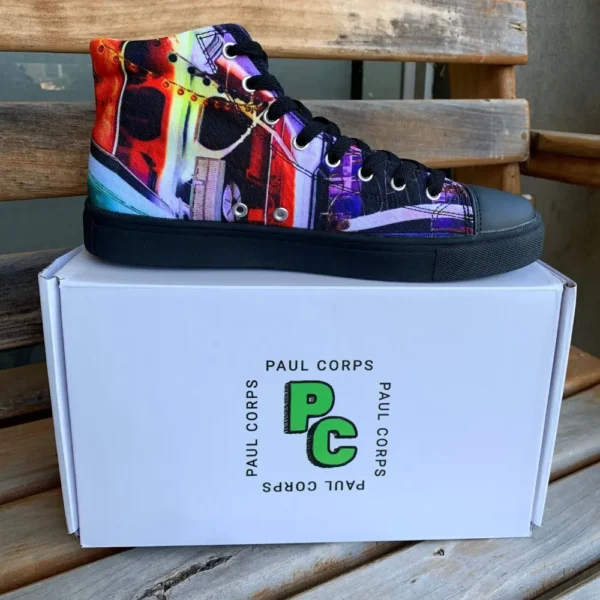 Paul Corps custom printed high-top canvas shoe featuring a vibrant, abstract cityscape design in bold colors with black laces and sole. With Shoe on top of Branded Paul Corps Shoe Box on wooden seat