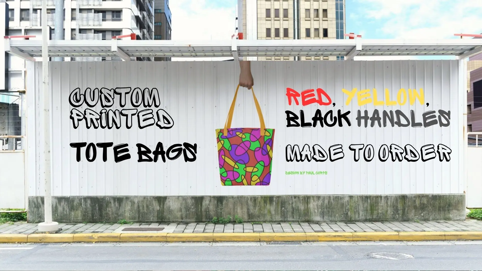 A street-side billboard displays a graphic advertisement for custom printed tote bags by Paul Corps, featuring a colorful abstract-patterned tote bag with yellow handles, held up by a hand, and bold graffiti-style text promoting made-to-order options with red, yellow, or black handles.