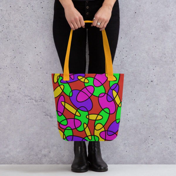 A person holding a Lava Lamp Memories all-over print tote bag by the yellow handles, featuring a bold abstract pattern in green, purple, yellow, and pink on a red background, standing against a light speckled wall