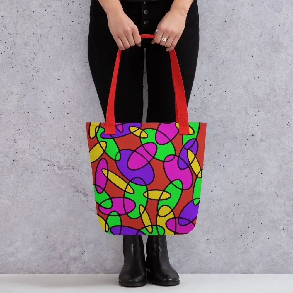 A person holding a Lava Lamp Memories all-over print tote bag by the red handles, featuring a bold abstract pattern in green, purple, yellow, and pink on a red background, standing against a light speckled wall