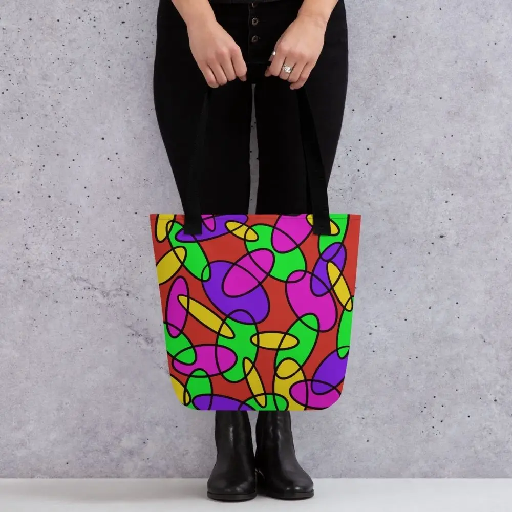 A person holding a Lava Lamp Memories all-over print tote bag by the black handles, featuring a bold abstract pattern in green, purple, yellow, and pink on a red background, standing against a light speckled wall
