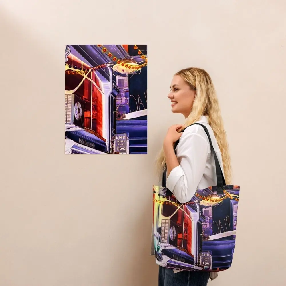 A Paul Corps tote bag featuring a vibrant, digitally enhanced photograph of a Melbourne laneway. The design includes colorful string lights, urban signage, and architectural details in shades of purple, orange, and blue, with black handles.
