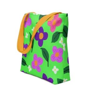 A green Spring all-over print tote bag with a vibrant flower and leaf design in pink, yellow, white and purple, set against a green background, featuring yellow handles