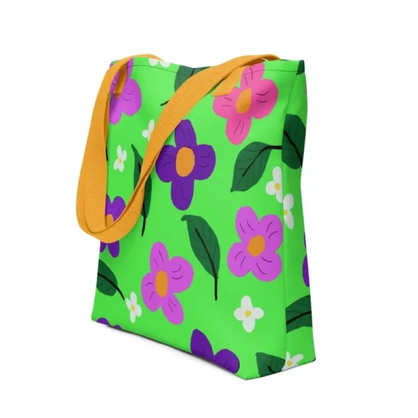 A green Spring all-over print tote bag with a vibrant flower and leaf design in pink, yellow, white and purple, set against a green background, featuring yellow handles