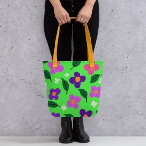 A person holding a Green Spring all-over print tote bag by the yellow handles, featuring a bold flower and leaf design in pink, yellow, white and purple, set against a green background standing against a light speckled wall