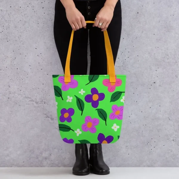 A person holding a Green Spring all-over print tote bag by the yellow handles, featuring a bold flower and leaf design in pink, yellow, white and purple, set against a green background standing against a light speckled wall