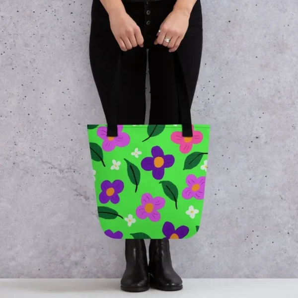 A person holding a Green Spring all-over print tote bag by the black handles, featuring a bold flower and leaf design in pink, yellow, white and purple, set against a green background standing against a light speckled wall