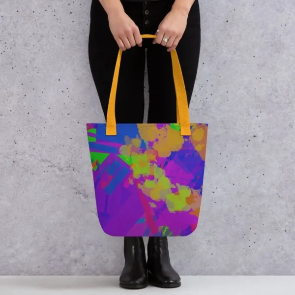 A person holding a Kaleidoscope Dream all-over print tote bag by the yellow handles, featuring a bold abstract pattern in green, purple, yellow, blue and pink, standing against a light speckled wall