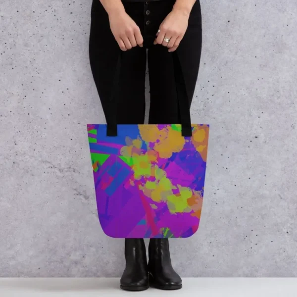 A person holding a Kaleidoscope Dream all-over print tote bag by the black handles, featuring a bold abstract pattern in green, purple, yellow, blue and pink, standing against a light speckled wall