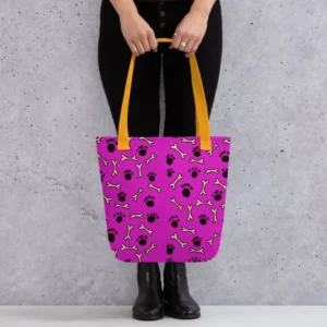 A Paul Corps large tote bag featuring a playful design of black paw prints and white bone motifs on a vibrant pink background, finished with sturdy black, red, yellow handles.