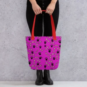 A Paul Corps large tote bag featuring a playful design of black paw prints and white bone motifs on a vibrant pink background, finished with sturdy black, red, yellow handles.