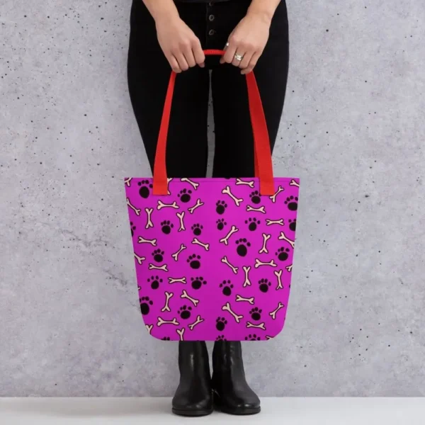 A Paul Corps large tote bag featuring a playful design of black paw prints and white bone motifs on a vibrant pink background, finished with sturdy black, red, yellow handles.