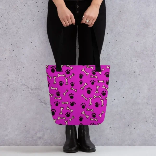 A Paul Corps large tote bag featuring a playful design of black paw prints and white bone motifs on a vibrant pink background, finished with sturdy black, red, yellow handles.