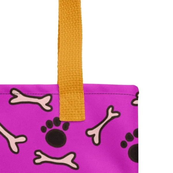 A Paul Corps large tote bag featuring a playful design of black paw prints and white bone motifs on a vibrant pink background, finished with sturdy black, red, yellow handles.