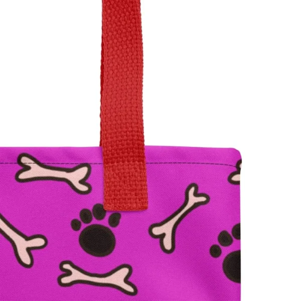 A Paul Corps large tote bag featuring a playful design of black paw prints and white bone motifs on a vibrant pink background, finished with sturdy black, red, yellow handles.