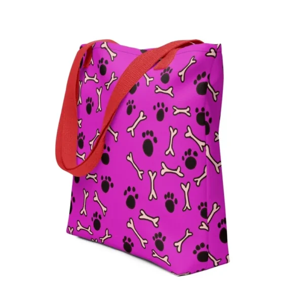 A Paul Corps large tote bag featuring a playful design of black paw prints and white bone motifs on a vibrant pink background, finished with sturdy black, red, yellow handles.
