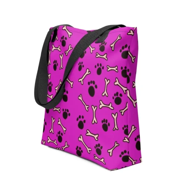 A Paul Corps large tote bag featuring a playful design of black paw prints and white bone motifs on a vibrant pink background, finished with sturdy black, red, yellow handles.