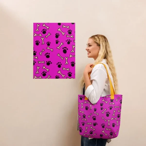 A Paul Corps large tote bag featuring a playful design of black paw prints and white bone motifs on a vibrant pink background, finished with sturdy black, red, yellow handles.