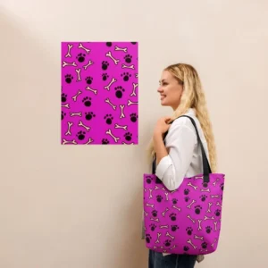 A Paul Corps large tote bag featuring a playful design of black paw prints and white bone motifs on a vibrant pink background, finished with sturdy black, red, yellow handles..