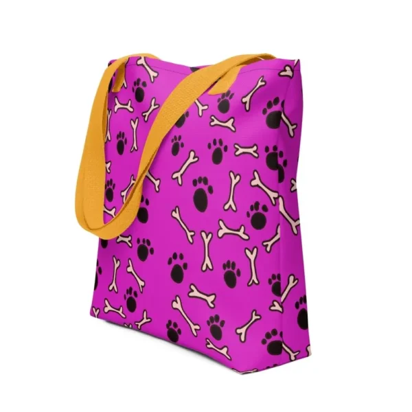 A Paul Corps large tote bag featuring a playful design of black paw prints and white bone motifs on a vibrant pink background, finished with sturdy black, red, yellow handles.
