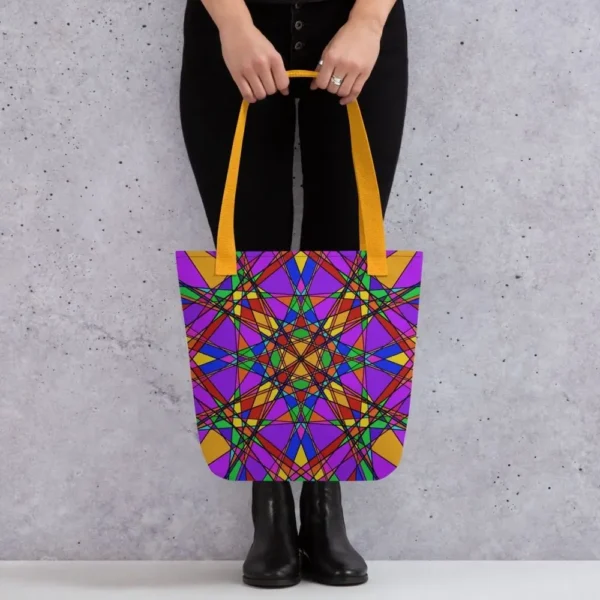 A person holding a Stain Glass Effect all-over print tote bag by the yellow handles, featuring a bold abstract pattern in green, purple, yellow, and pink, standing against a light speckled wall