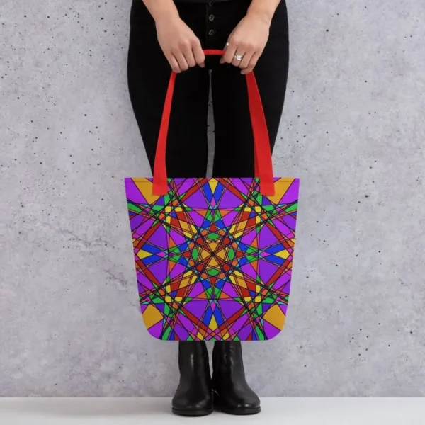 A person holding a Stain Glass Effect all-over print tote bag by the red handles, featuring a bold abstract pattern in green, purple, yellow, and pink, standing against a light speckled wall