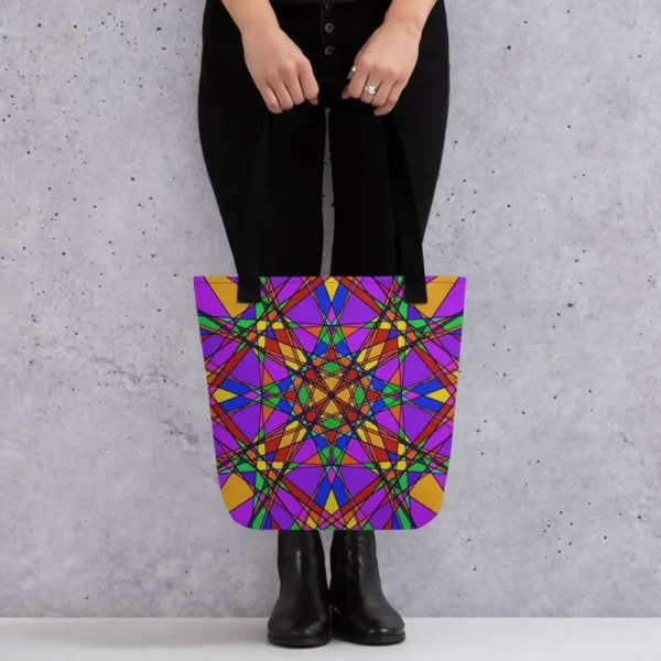 A person holding a Stain Glass Effect all-over print tote bag by the black handles, featuring a bold abstract pattern in green, purple, yellow, and pink, standing against a light speckled wall